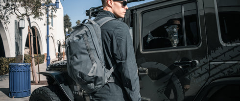 A person wearing sunglasses and a Condor Elite Fail Safe Urban Backpack Gen II 18L by Condor stands next to a dark off-road vehicle on a sunny street.